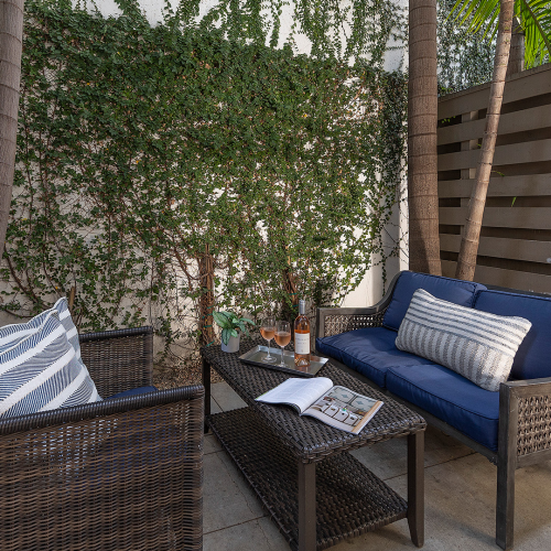 A cozy outdoor seating area with wicker furniture, blue cushions, striped pillows, a glass coffee table, surrounded by trees and a wooden fence.