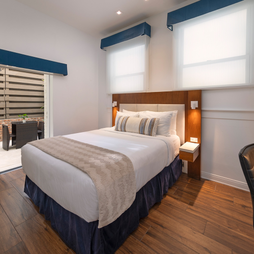 A modern hotel room with a king bed, two nightstands, desk and chair, wooden floor, white walls, blue valances, and a doorway to a balcony or seating area.