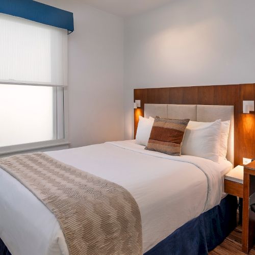 A tidy hotel bedroom with a queen bed, wooden headboard, two bedside lamps, a wall-mounted TV, a window with a shade, and a small desk with chair.