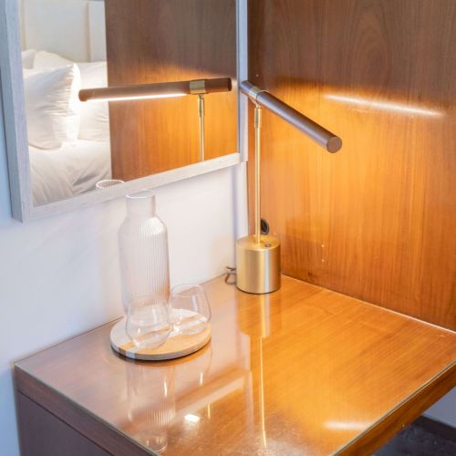A wooden desk with a glass carafe, matching water glass, and a modern wall-mounted lamp casting light on the surface, near a mirror.