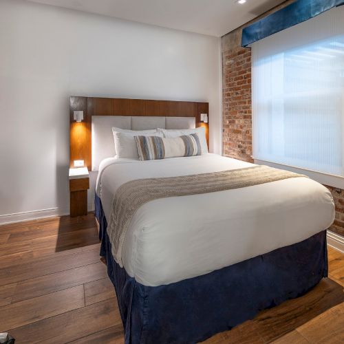 A modern hotel room with a queen bed, brick accent wall, wooden floor, desk and chair, wall-mounted TV, bedside lamps, and a large window.