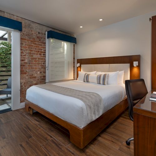 A contemporary hotel room with a wooden bed, desk, and chair; glass doors open to a small outdoor patio with a plant and seating.