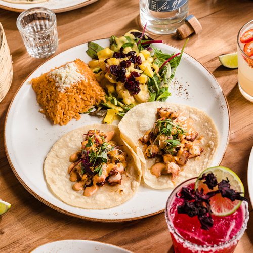 A plate with two tortillas and toppings, creamy sauces, rice, beans, a mixed salad, salsa, a mango drink, and a pink smoothie on a wooden table.