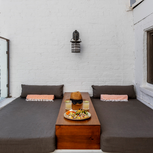 An outdoor lounge area with two gray cushioned benches flanking a wooden center table, topped by a pizza and drinks on a sunny white brick patio.