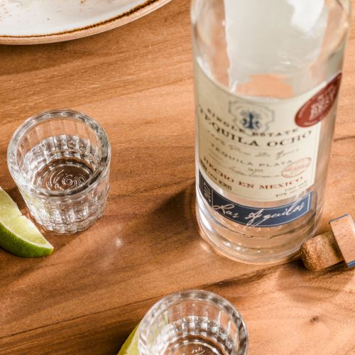A bottle of liquor with a label, a shot glass, and a small lime wedge on a wooden table.