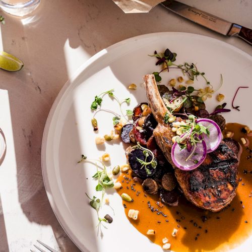 A plated gourmet dish with a sauce, grilled meat, onion slices, microgreens, and a side of chips; sunlit table setting and a drink nearby.