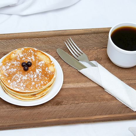 A wooden tray with a stack of pancakes topped with berries, a cup of black coffee, and a neatly folded napkin with a fork on a white cloth backdrop.