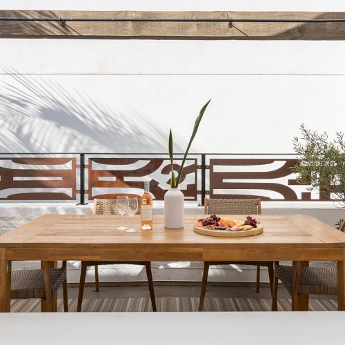 A cozy kitchen with a wooden dining table, chairs, and a breakfast bar; warm light, potted plants, and a window view outside.