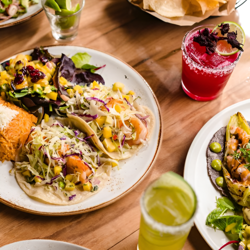 A lively spread: tacos with cabbage slaw, tortillas, a red drink with lime, a green drink, and small bites on a wooden table.