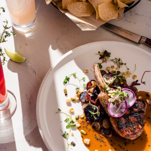 A plated dish with grilled meat, onions, greens, and a drizzle of sauce, plus tortilla chips and lime nearby, on a bright table.