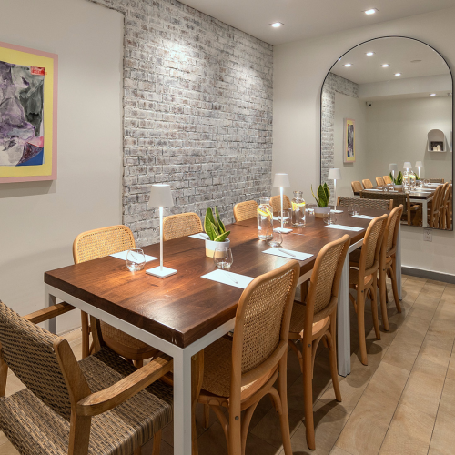 A cozy, narrow dining nook with a wooden table set for two, warm lighting, decorative mirror, and soft wall art lining a small hallway.