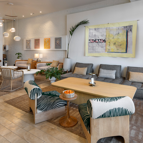 A bright, cozy living room with a textured rug, light wood floors, mustard sofas, a glass coffee table, a large wall art and potted plants, inviting ambiance.