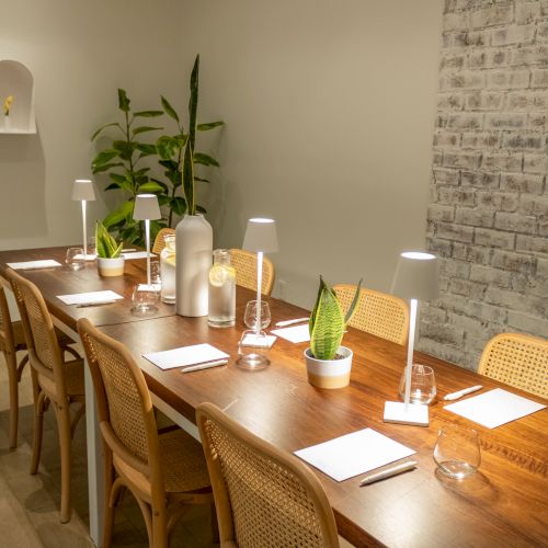 A cozy dining table set for a meal in a rustic room with brick wall, plants, candles, and warm lighting, ready for guests.