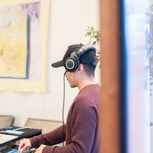 A young man wearing headphones sits at a desk with a laptop and DJ gear, mixing music in a cozy room with artwork on the wall.