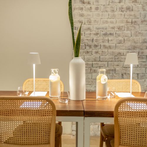 A minimal dining setup with a tall plant vase, scented bottles, and warm lamps around a wooden table, wicker chairs, and a brick wall.