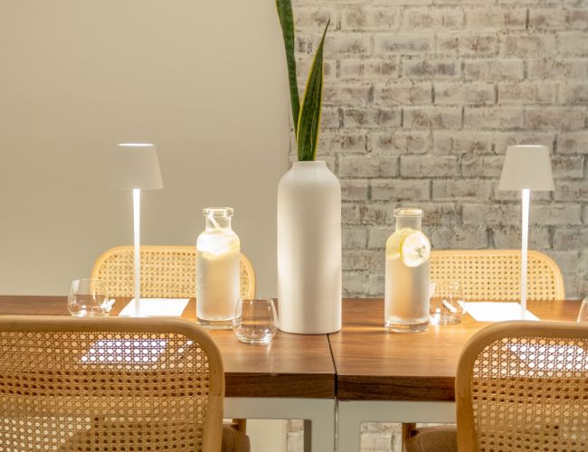 A minimal dining setup with a tall plant vase, scented bottles, and warm lamps around a wooden table, wicker chairs, and a brick wall.