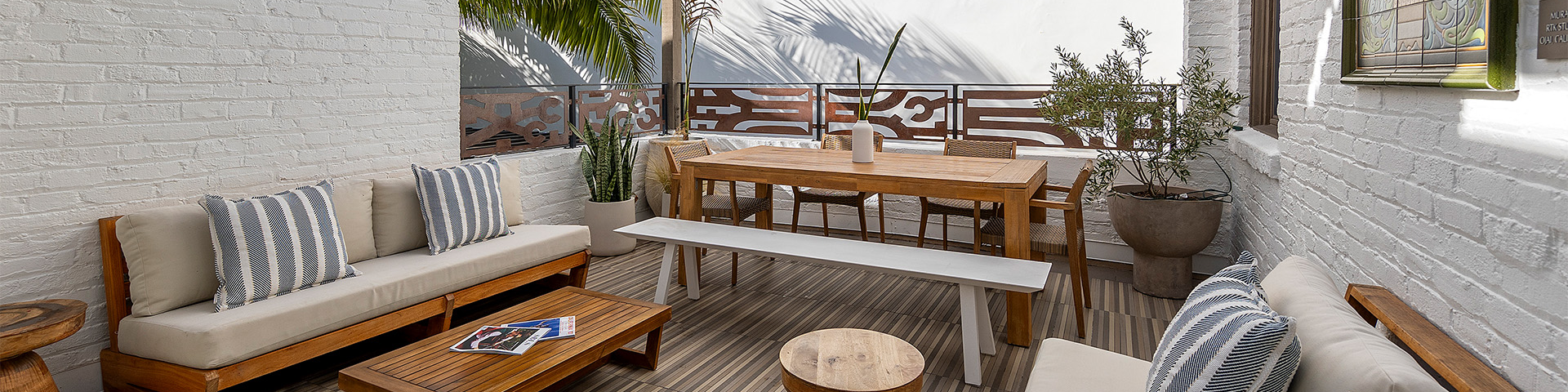 A cozy patio with white brick walls, wooden furniture, striped cushions, potted plants, and a shaded dining area.