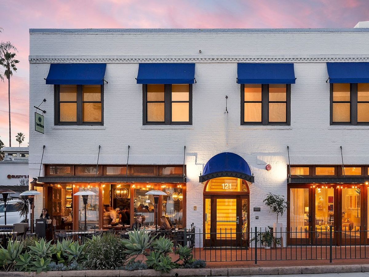 A charming two-story white building with blue awnings, warm-lit windows, outdoor seating, and palm trees, glowing at dusk.
