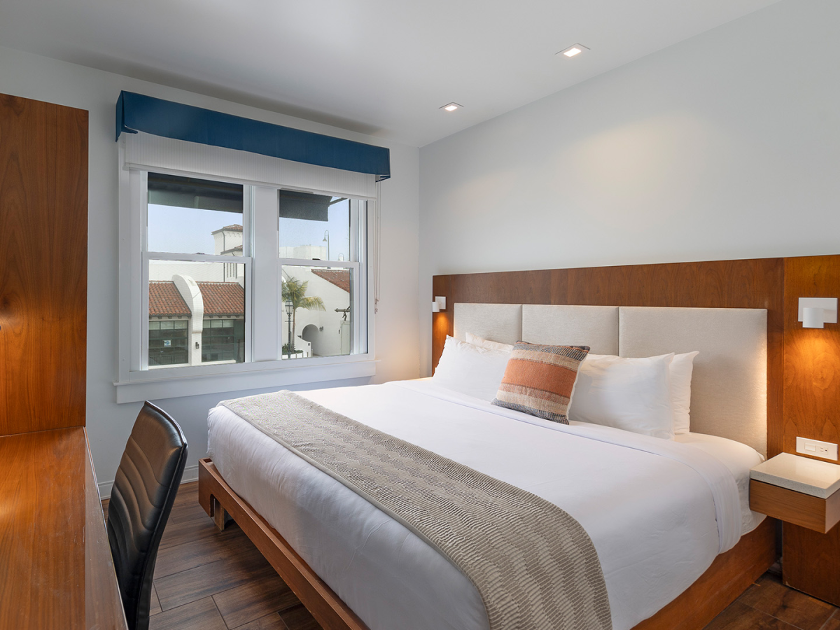 A modern hotel room with a large bed, wooden headboard, bedside lamps, a desk with a chair, and a window letting in natural light.