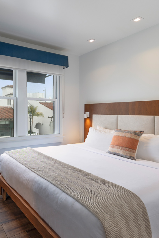 A modern hotel room with a large bed, wooden headboard, bedside lamps, a desk with a chair, and a window letting in natural light.