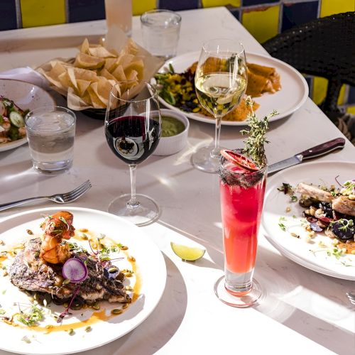 A fine-dining table with plated entrees, wine glasses, and a colorful pink cocktail in bright light, ready to enjoy.