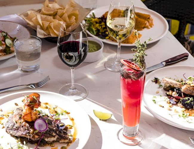 A fine-dining table with plated entrees, wine glasses, and a colorful pink cocktail in bright light, ready to enjoy.