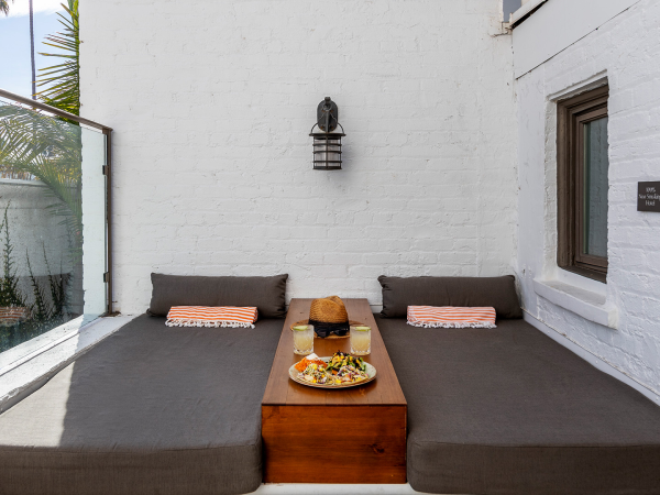 A sunny patio with gray cushions, striped pillows, a wooden table, drinks and a salad, framed by palm trees.