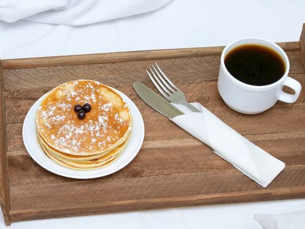 A wooden tray holds a stack of pancakes with blueberries, a fork and knife, and a cup of black coffee.