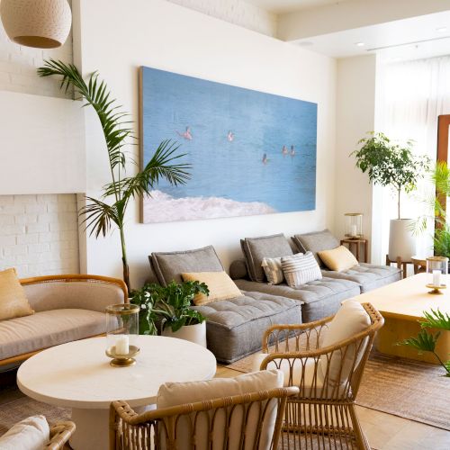 A bright modern living room with a gray sectional, round coffee table, plants, and a large ocean-themed artwork on the wall.