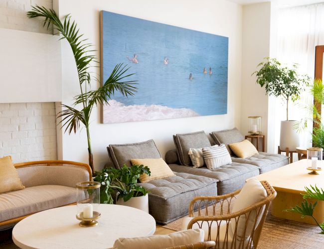 A bright modern living room with a gray sectional, round coffee table, plants, and a large ocean-themed artwork on the wall.