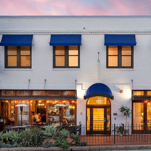 A white, two-story building with blue awnings, a lit entrance marked &ldquo;121,&rdquo; and a restaurant patio with people dining at sunset.