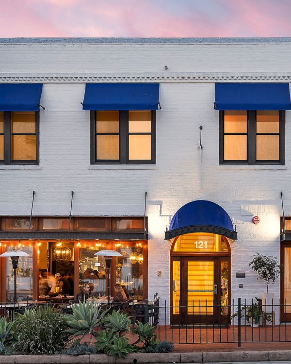 A white, two-story building with blue awnings, a lit entrance marked &ldquo;121,&rdquo; and a restaurant patio with people dining at sunset.