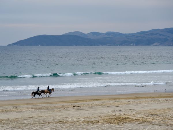 A calm beach with waves and a couple riding horses along the shoreline while distant hills fade into the horizon.