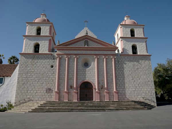 A white stone church with twin bell towers, pink columns, and a cross on the domes against a blue sky.