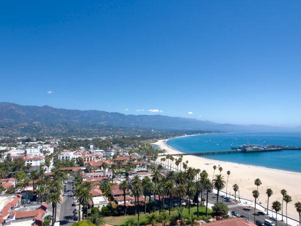 Coastal town with palm-lined houses, a wide sandy beach, blue ocean, and distant mountains under a clear sky.