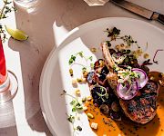 A plated entree: grilled chops over orange sauce with radish, microgreens, and herbs, next to a lime water bottle and chips.