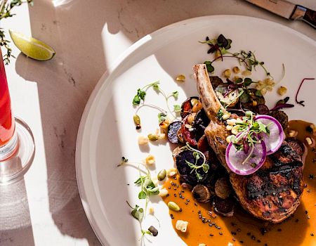 A plated entree: grilled chops over orange sauce with radish, microgreens, and herbs, next to a lime water bottle and chips.