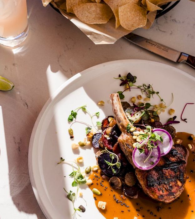 A plated entree: grilled chops over orange sauce with radish, microgreens, and herbs, next to a lime water bottle and chips.