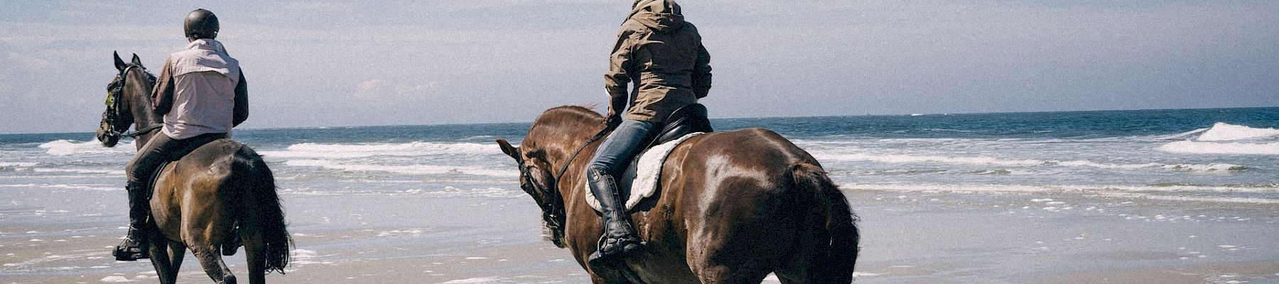 Two helmeted riders on horseback trot along a sandy beach with waves under a clear sky.