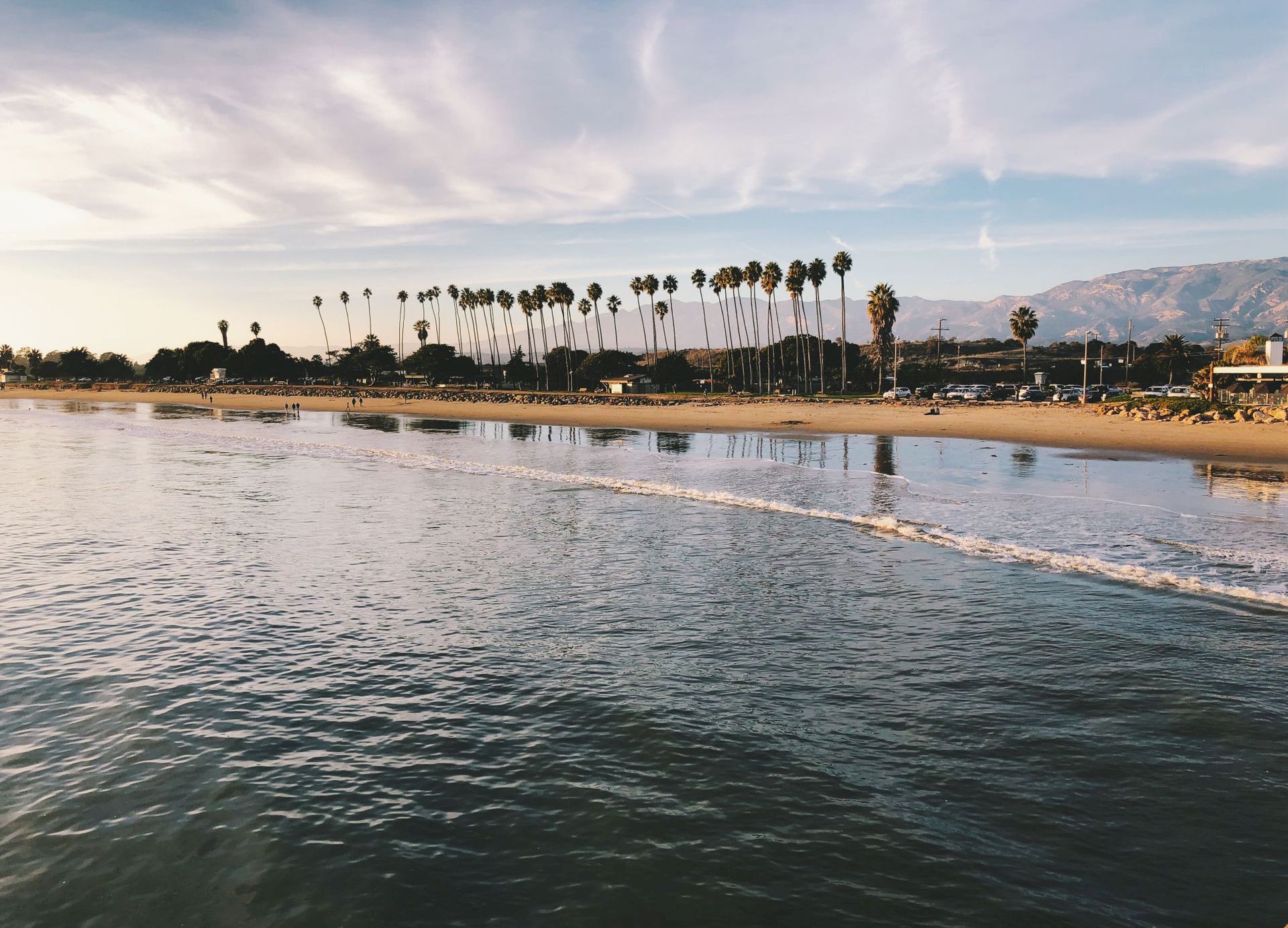 A calm beach at sunset with gentle waves, rows of tall palm trees, a distant coastline, and mountains under a cloudy sky.