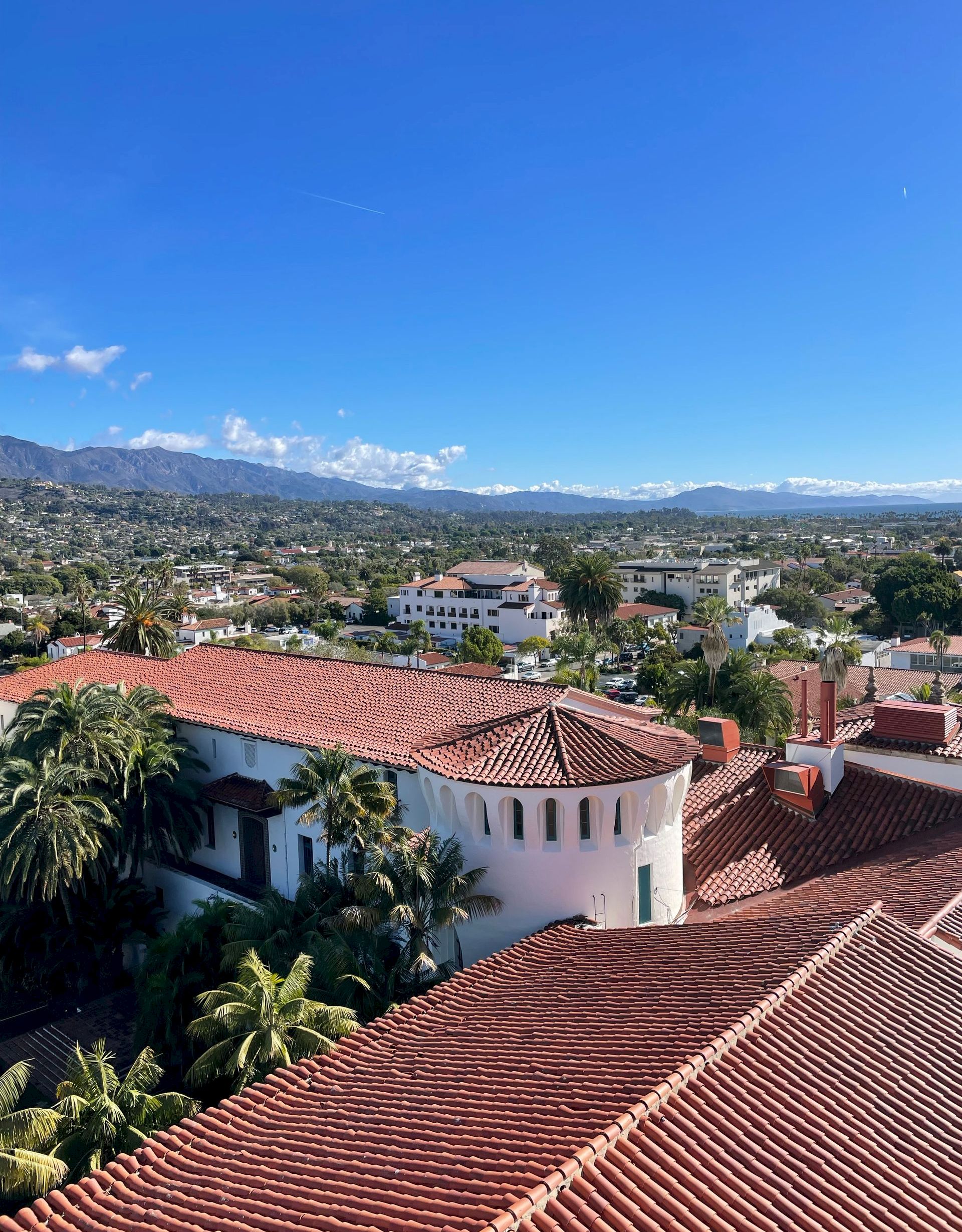 Sunlit hillside town with red-tile rooftops, palm trees, and distant mountains beneath a vast blue sky and clouds.