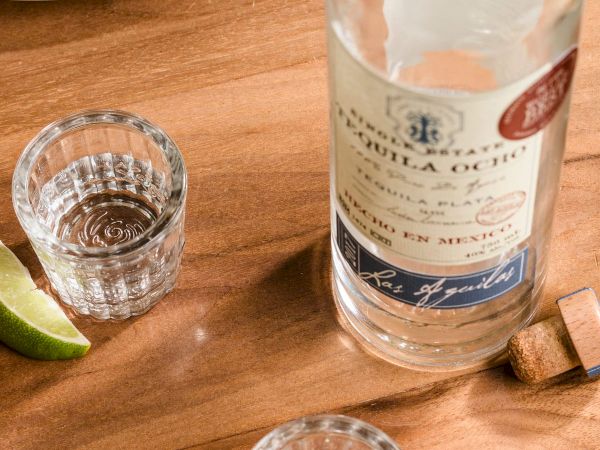 A taco plate sits beside a tequila bottle and shot glasses with lime wedges on a wooden table, ready to drink.