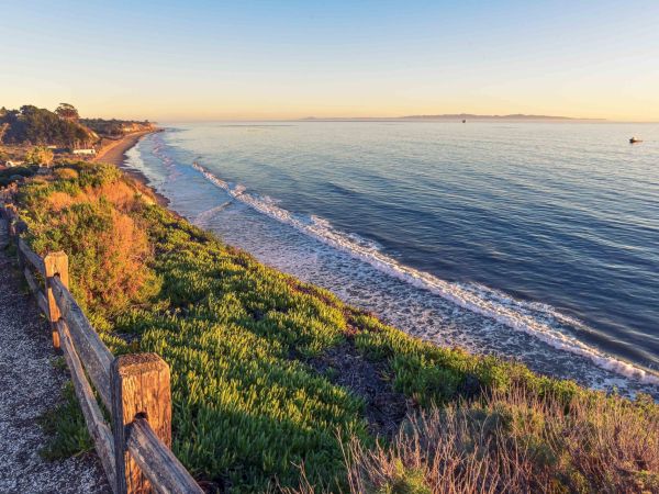 A scenic coastline at sunset with grassy cliffs, a wooden fence, and waves rolling in under a pastel sky.