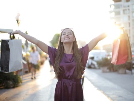 A smiling woman shopping on a sunny street, holding colorful bags outstretched in celebration.