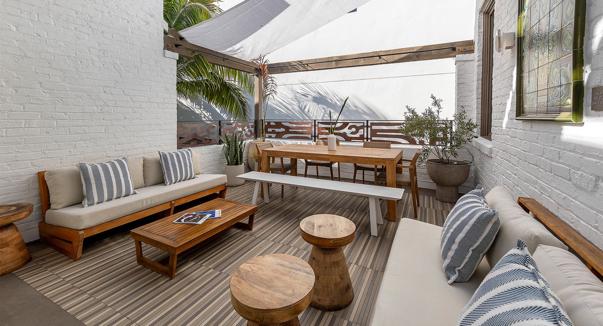 A bright outdoor patio with wood benches and tables, striped cushions, potted plants, and a shaded pergola.