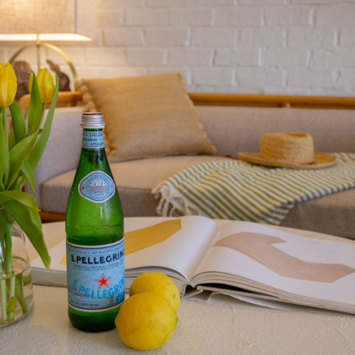 A tabletop scene with yellow tulips in a vase, an S. Pellegrino bottle, lemons, and an open book in a cozy lounge.