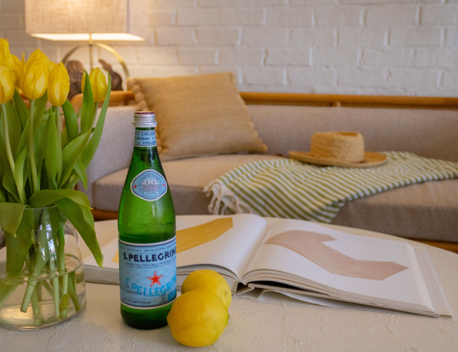 A tabletop scene with yellow tulips in a vase, an S. Pellegrino bottle, lemons, and an open book in a cozy lounge.