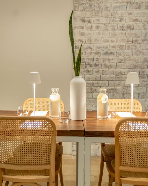 A dining table styled with a tall white vase of green leaves, amber bottles, two lamps, and a brick wall backdrop.