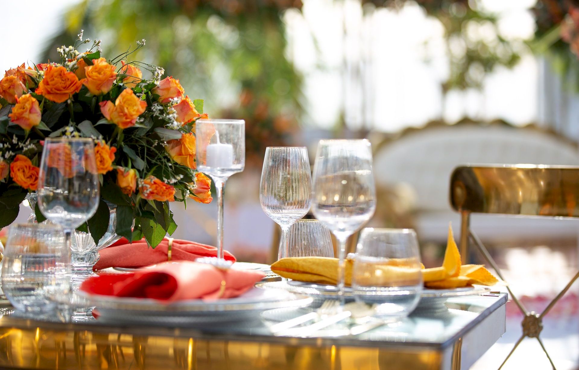 A beautifully set outdoor banquet table with orange roses, sparkling wine glasses, folded napkins, and a warm, golden ambiance.