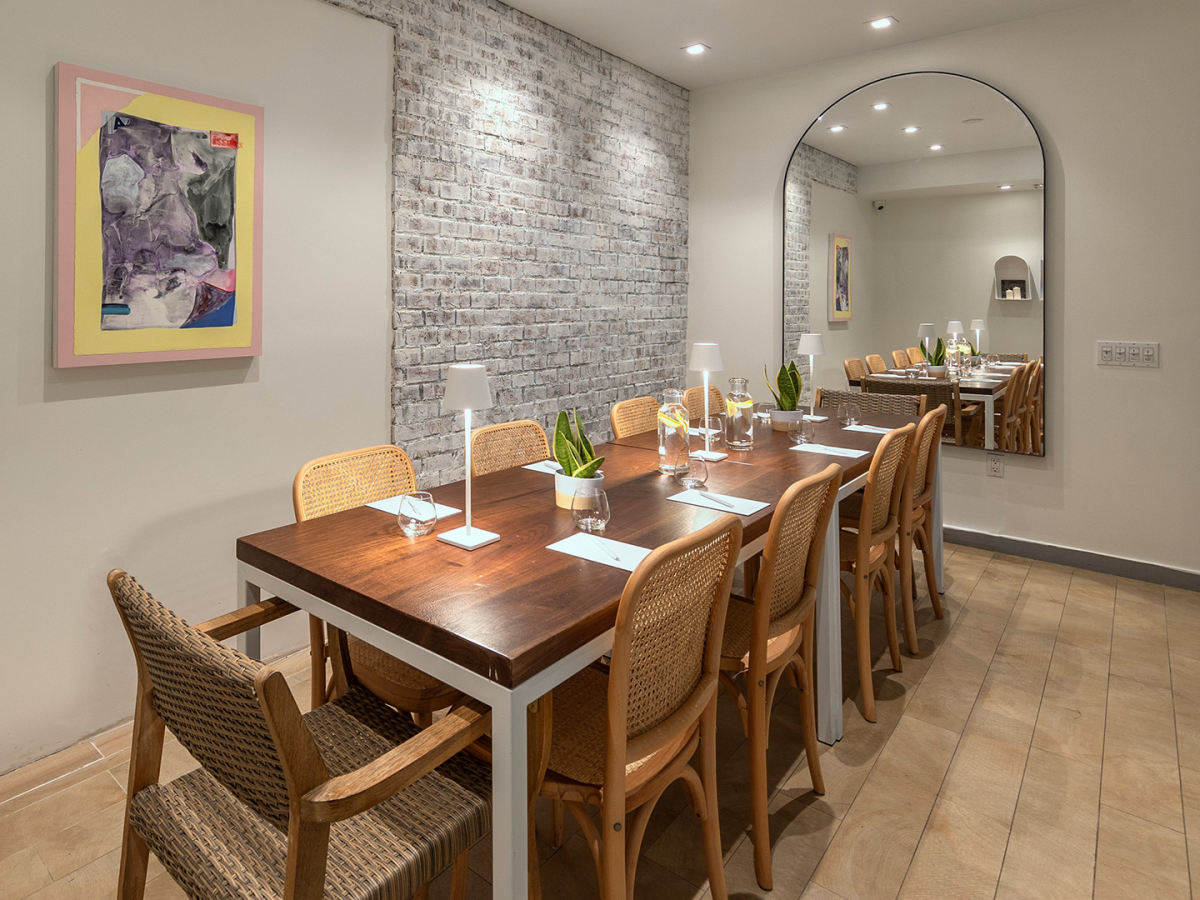 A modern dining room with a long wooden table, wicker chairs, brick wall, and framed artwork.